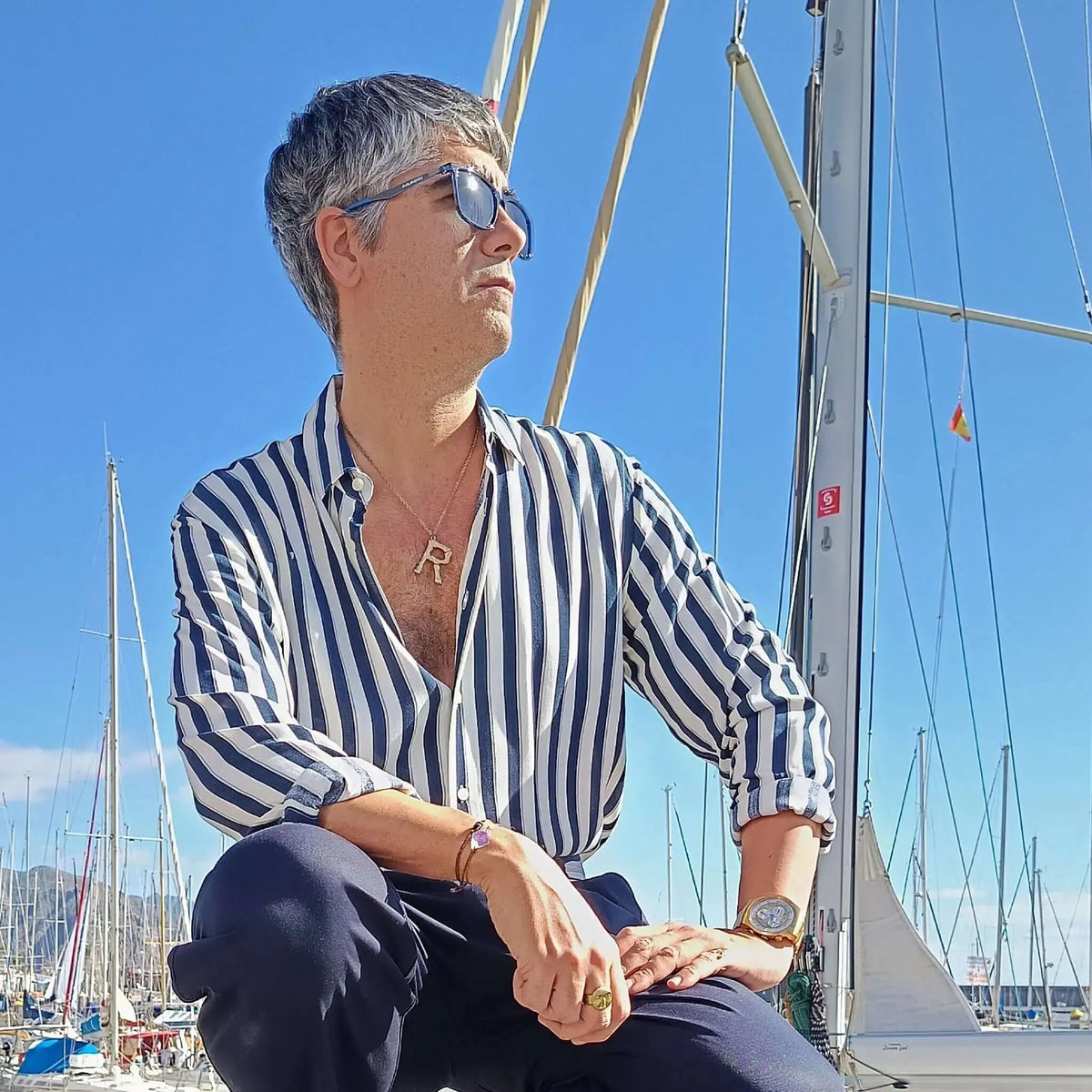 Hombre con camisa a rayas y gafas de sol de montura azul, posando frente a un velero en un puerto deportivo soleado.