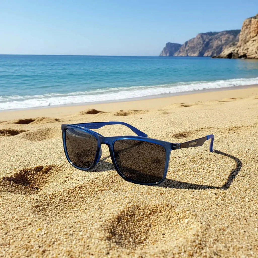 Gafas de sol con montura azul transparente y cristales oscuros sobre la arena de la playa con mar de fondo.