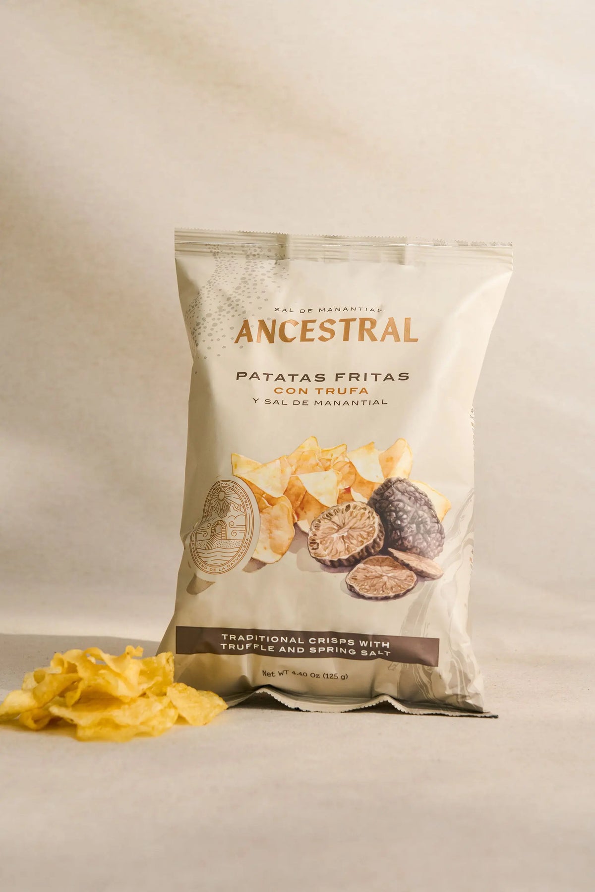 Bag of gourmet truffle and spring salt potato chips with a pile of loose chips on a beige background.