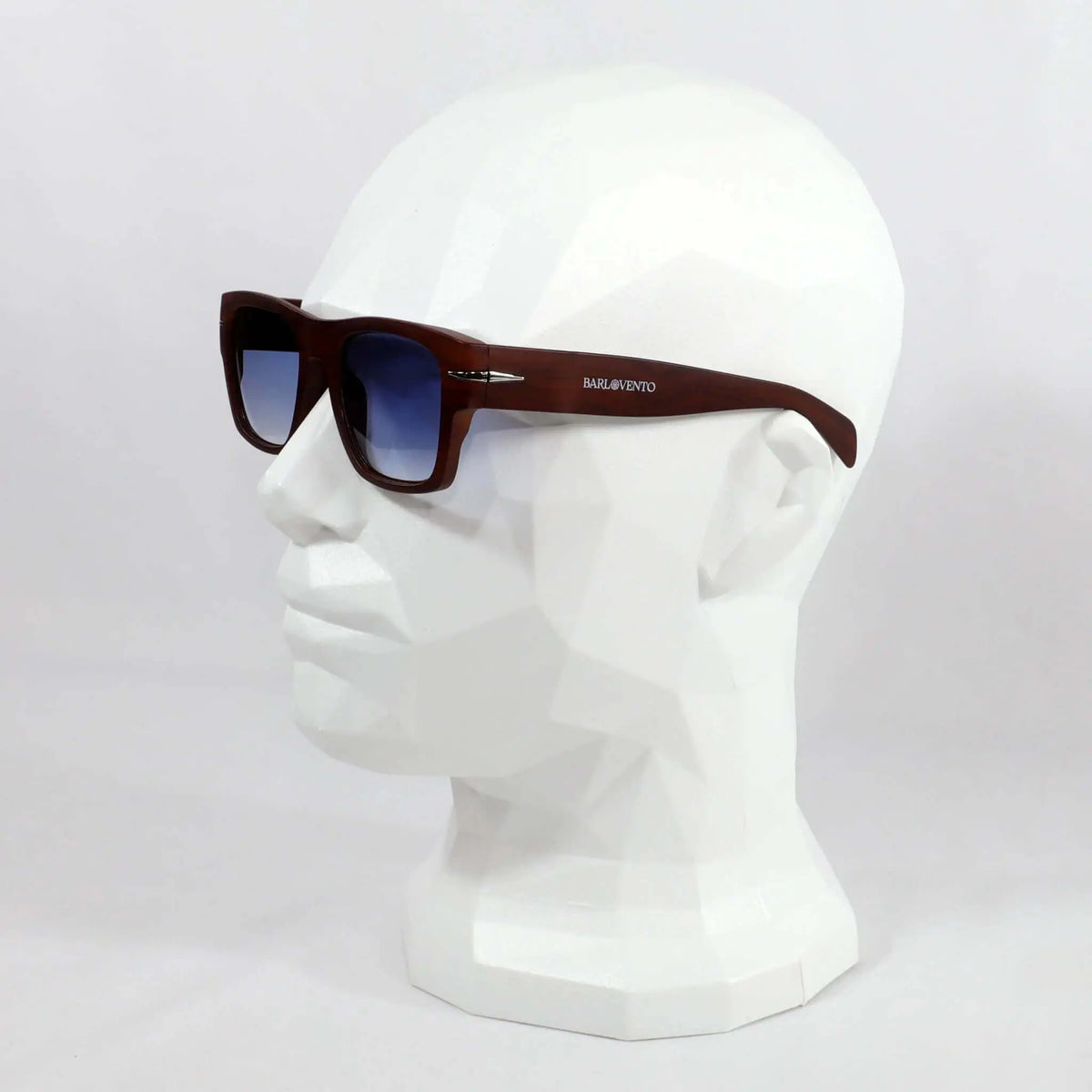 Gafas de sol cuadradas con montura marrón efecto madera y lentes azules degradadas sobre un busto blanco.
