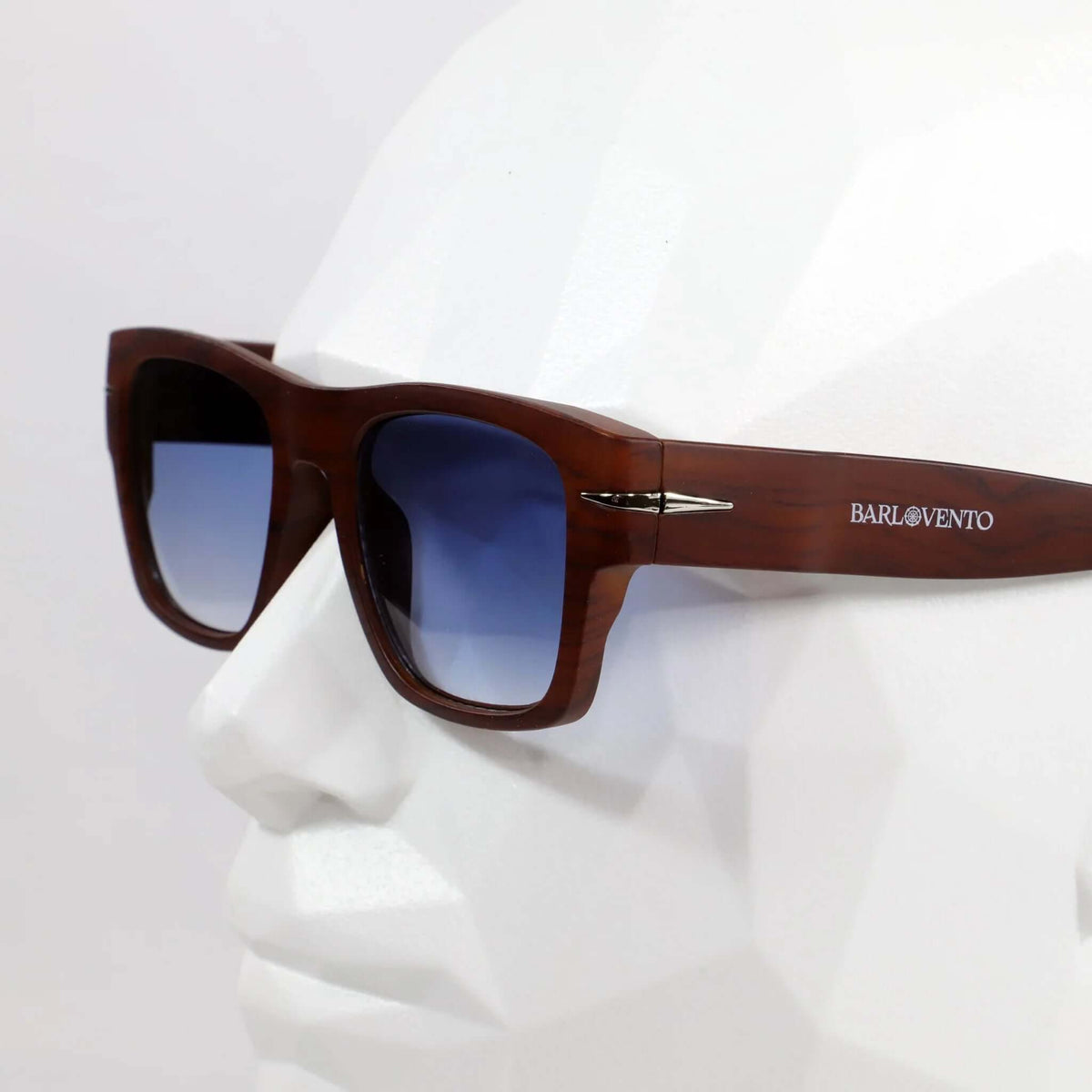 Gafas de sol cuadradas con montura estilo madera marrón y lentes degradadas azules puestas en un maniquí blanco.