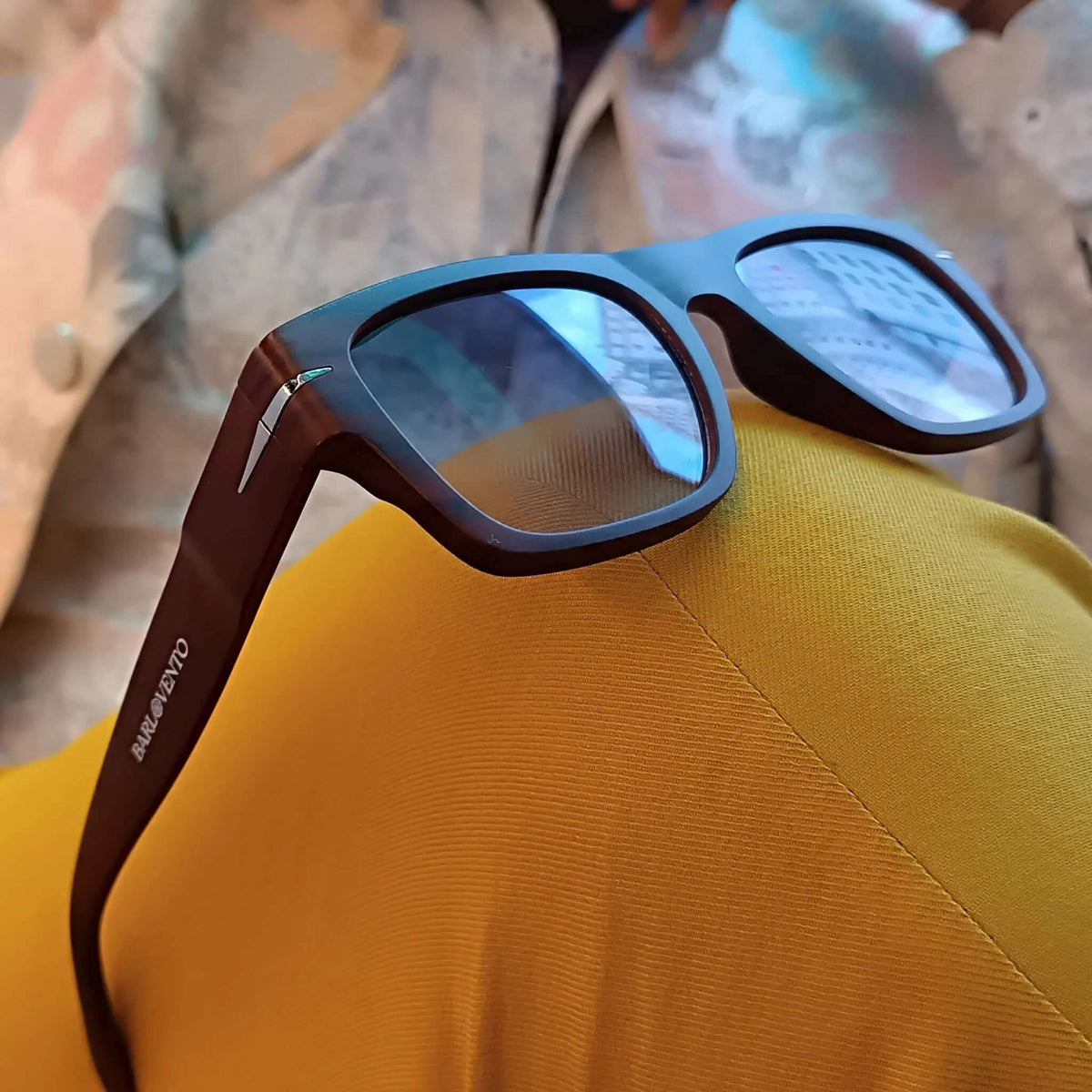 Gafas de sol cuadradas con montura de madera oscura y lentes azules sobre ropa de color amarillo.