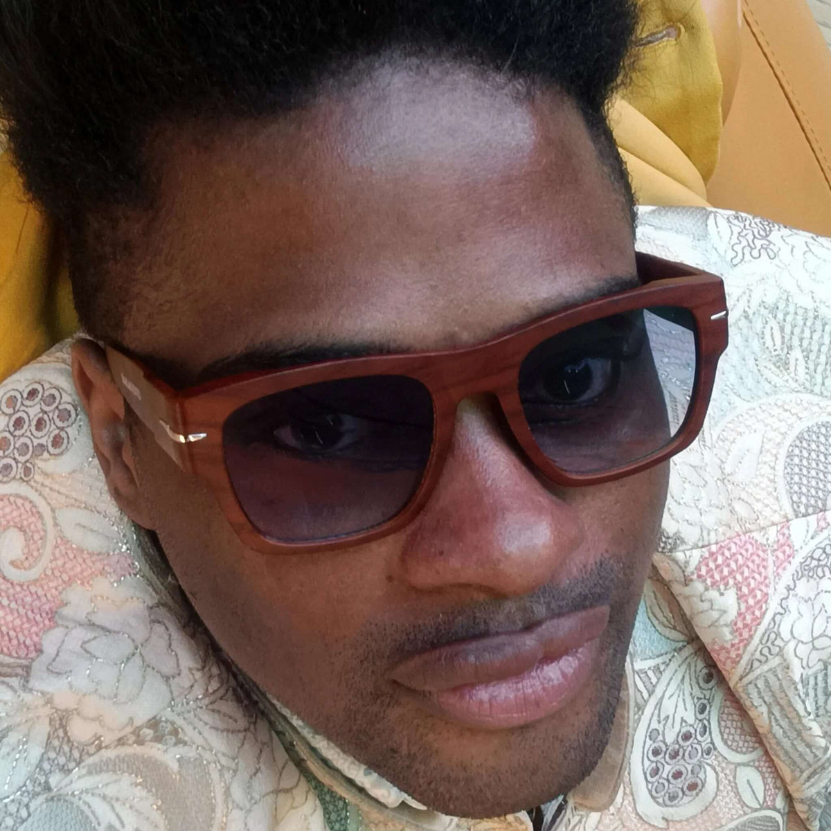 Hombre con gafas de sol cuadradas de madera marrón y lentes azules, vistiendo chaqueta estampada elegante.