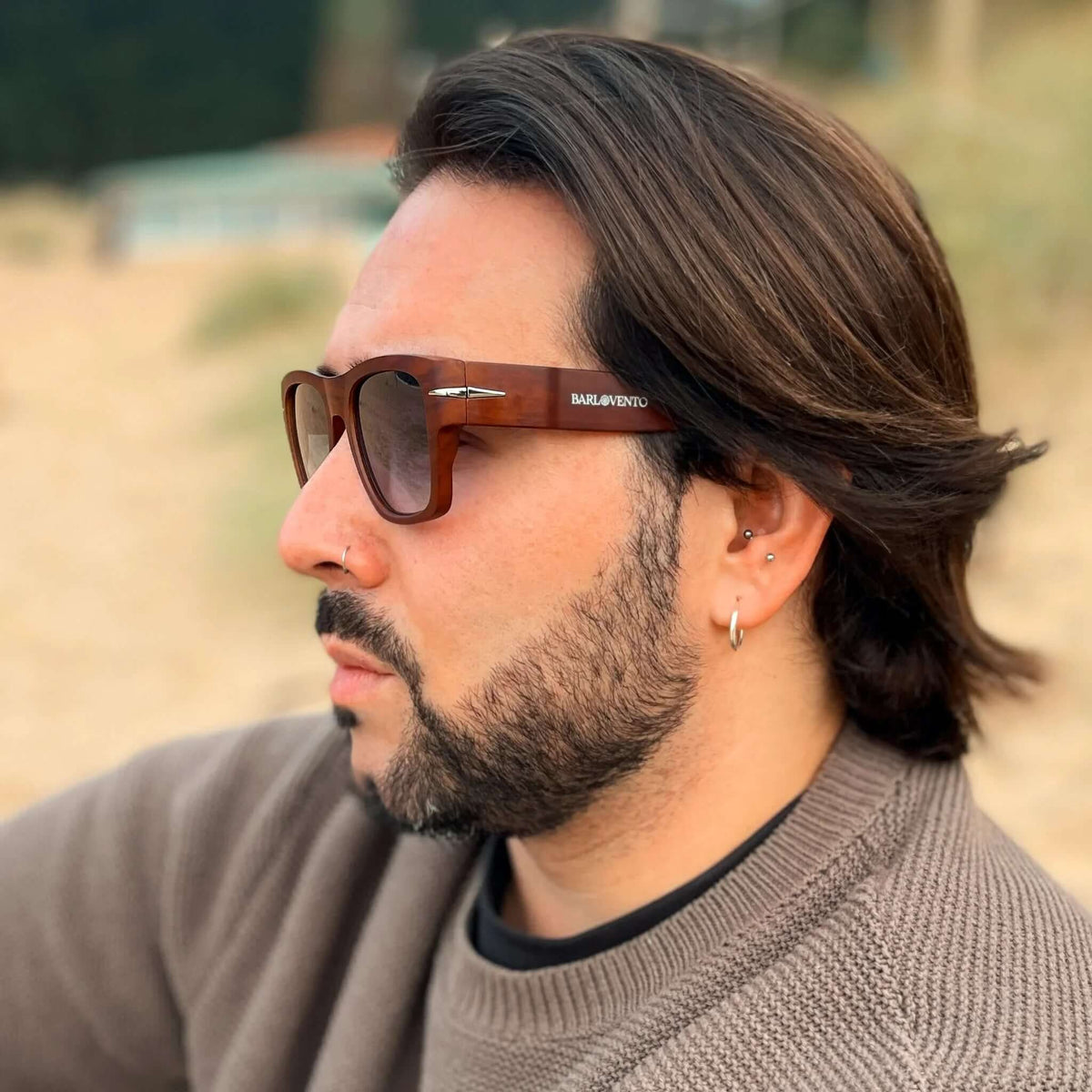 Hombre con barba y gafas de sol cuadradas con montura efecto madera y cristales oscuros en un entorno de playa.