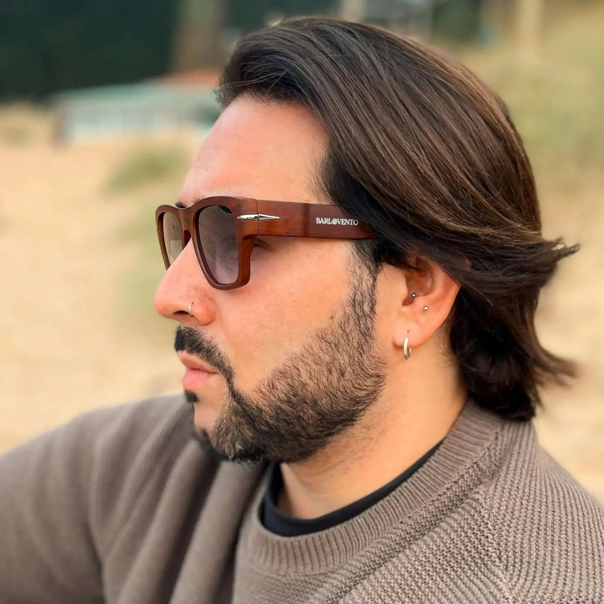 Hombre con barba y gafas de sol cuadradas con montura efecto madera y cristales oscuros en un entorno de playa.