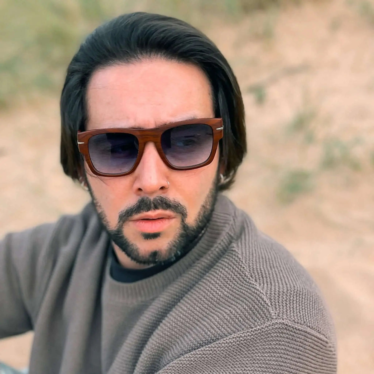 Hombre con suéter gris usando gafas de sol cuadradas con marco de madera marrón y lentes azules degradados en exterior.
