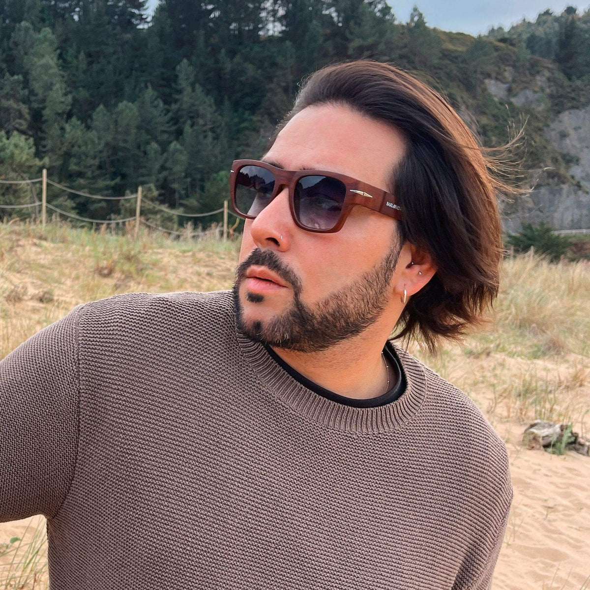 Hombre con gafas de sol cuadradas efecto madera y lentes azules oscuras, vistiendo suéter beige en paisaje costero.