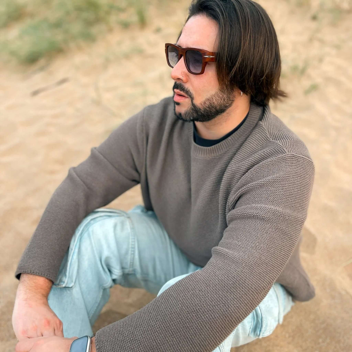 Hombre con gafas de sol cuadradas efecto madera y lentes azules sentado en la arena, usando suéter marrón y jeans.