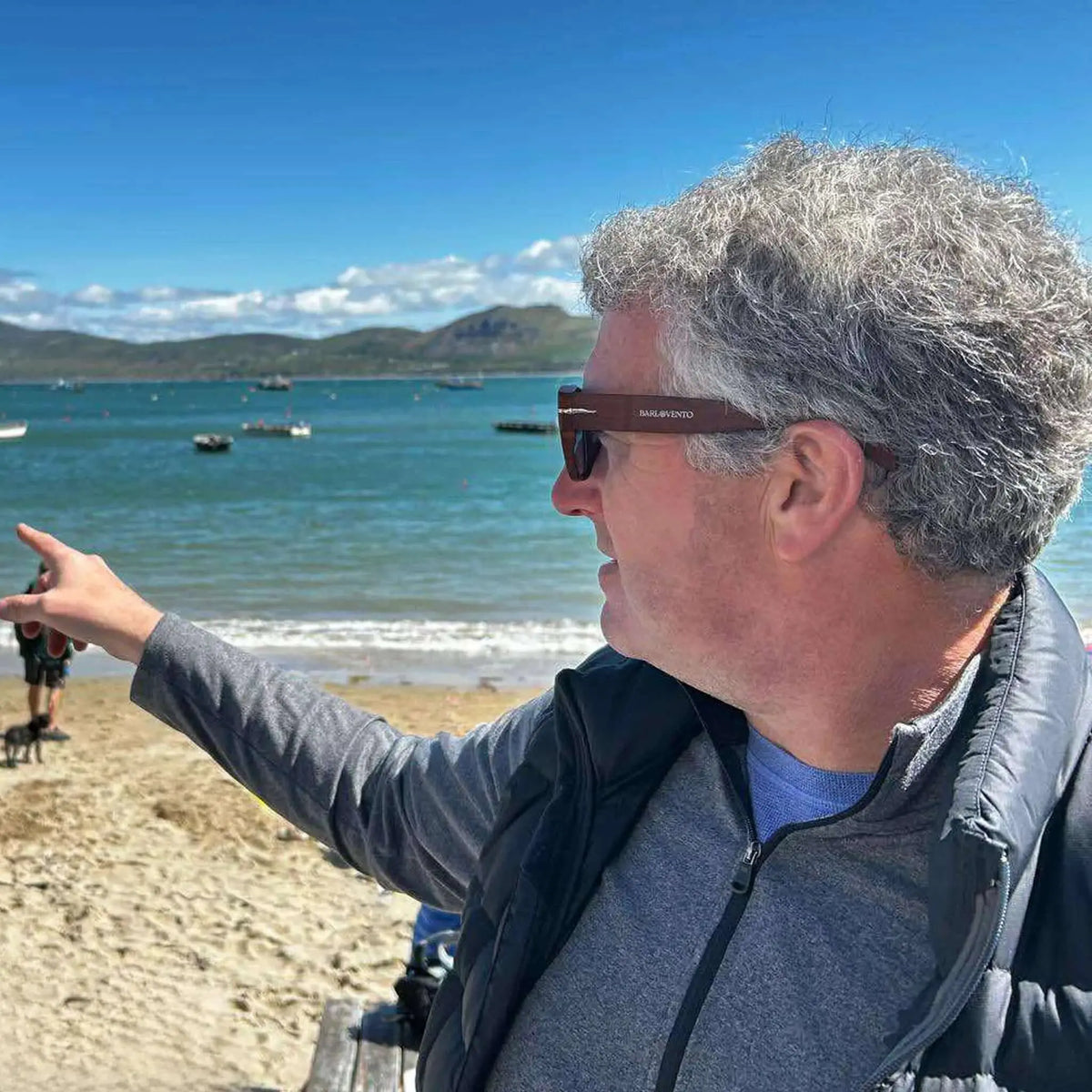 Hombre con gafas de sol cuadradas de madera y lentes azules señalando al mar en la playa.