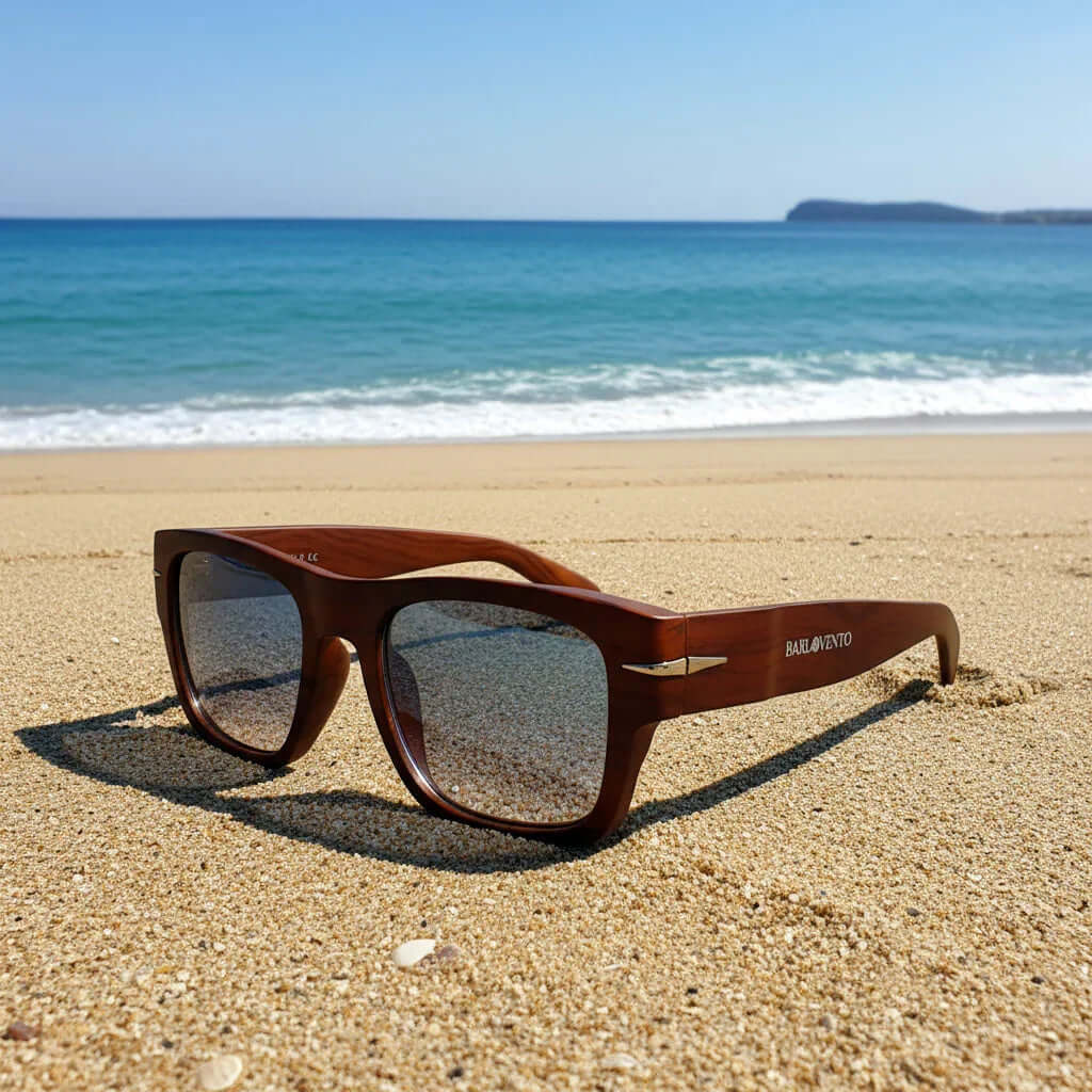 Gafas de sol cuadradas con montura de madera natural y lentes azules en la arena de una playa frente al mar.
