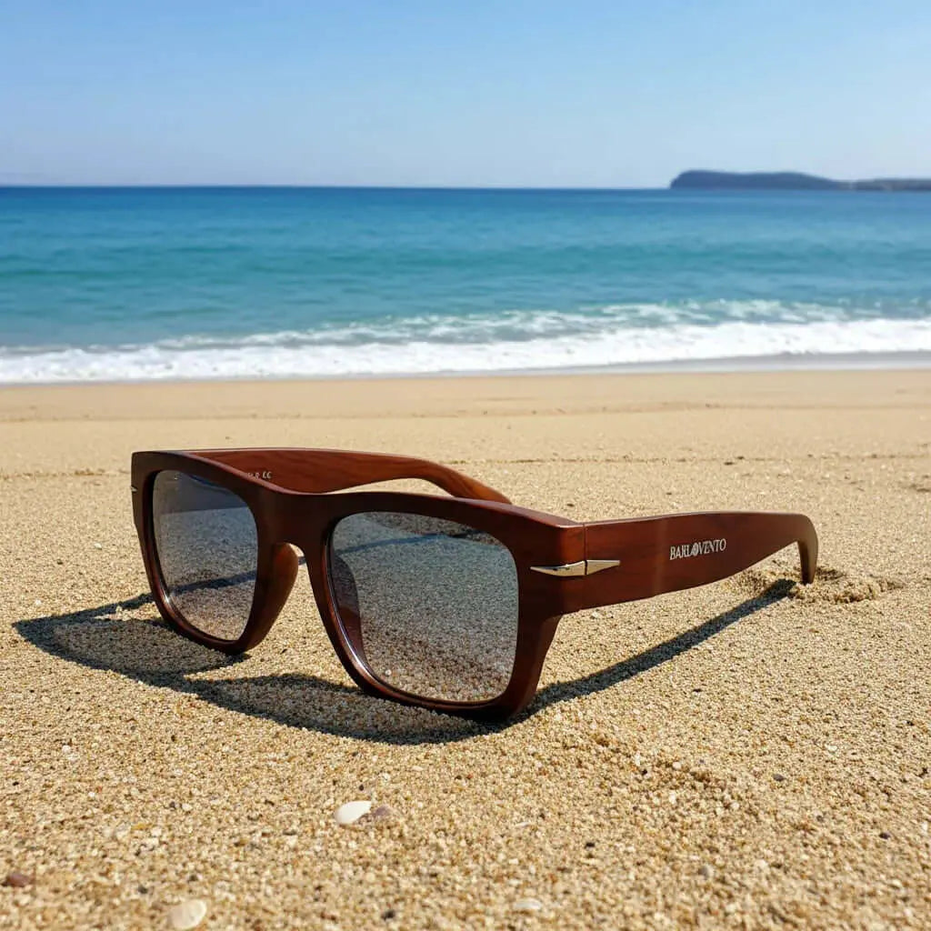 Gafas de sol cuadradas con montura de madera natural y lentes azules en la arena de una playa frente al mar.