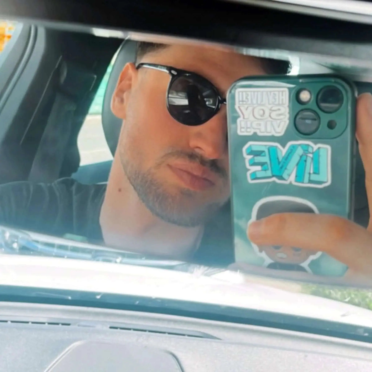 Hombre con gafas de sol negras de montura redondeada y lentes polarizadas verdes, reflejado en el espejo de un coche.