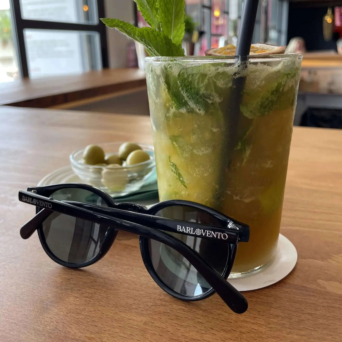 Gafas de sol negras de estilo clásico con lentes polarizadas verdes junto a un cóctel refrescante en una mesa.