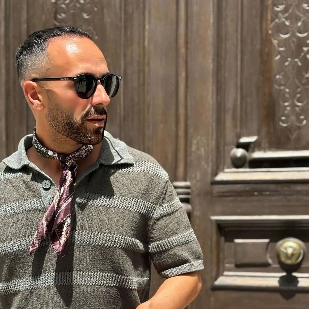 Hombre con gafas de sol redondas de pasta negra y lentes verdes polarizadas, estilo casual ante una puerta de madera.