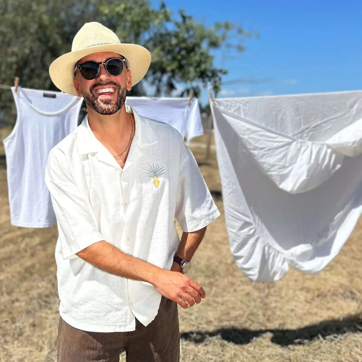 Hombre con sombrero, camisa blanca y gafas de sol negras de montura redonda con lentes polarizadas verdes al aire libre.