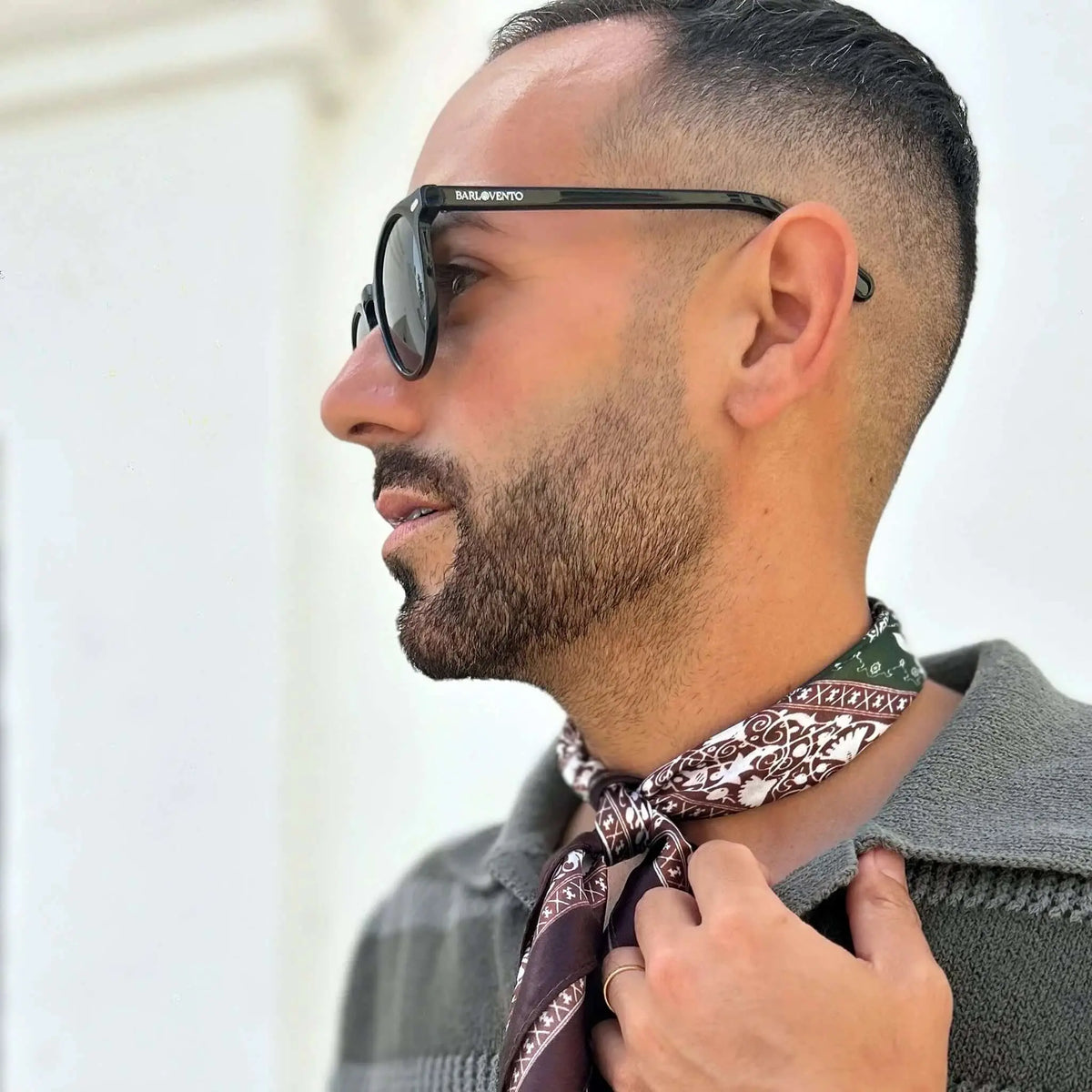 Hombre con gafas de sol negras de montura clásica y lentes polarizadas verdes, usando pañuelo estampado y polo gris.
