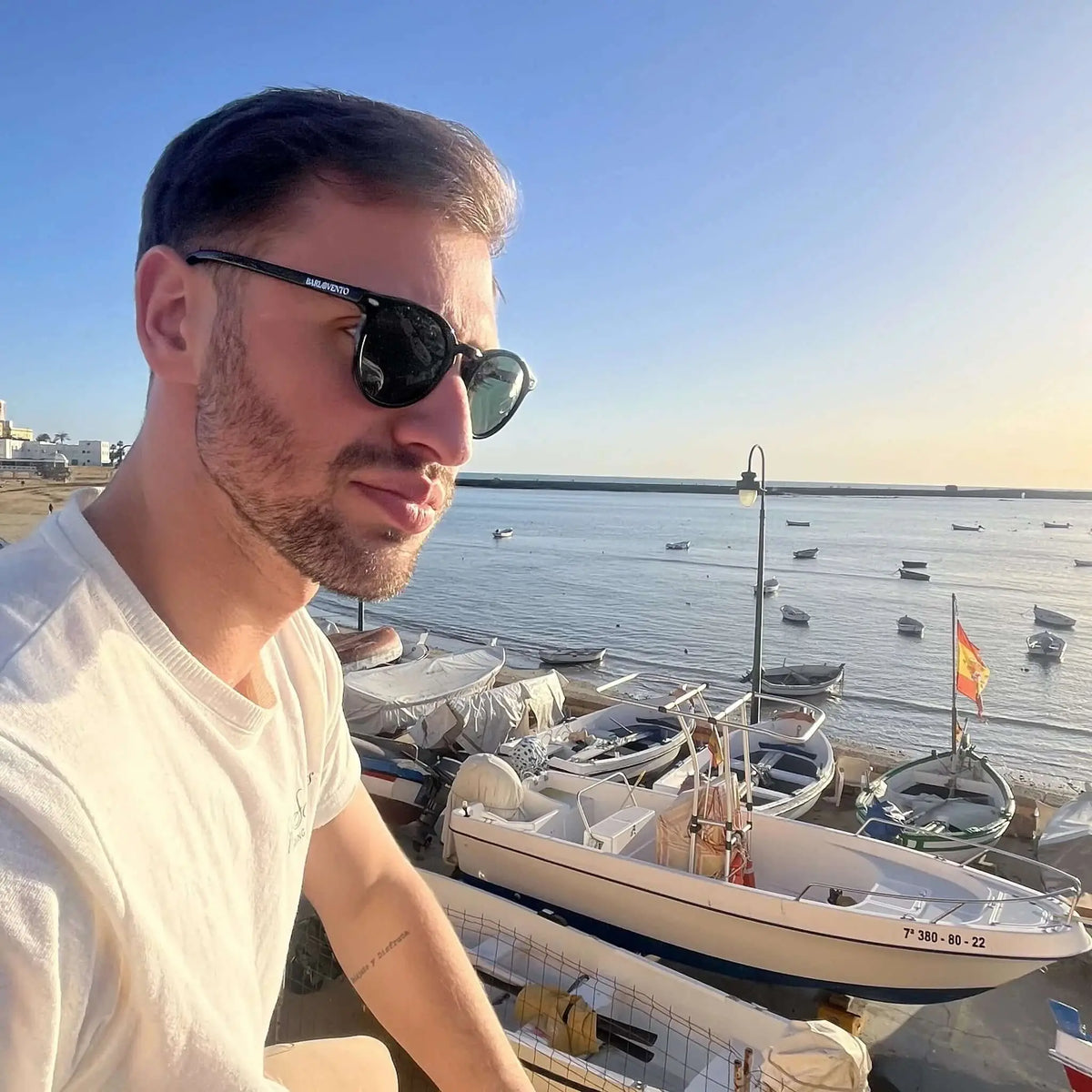 Hombre con camiseta blanca usa gafas de sol negras con lentes polarizadas verdes frente a un puerto con barcos.