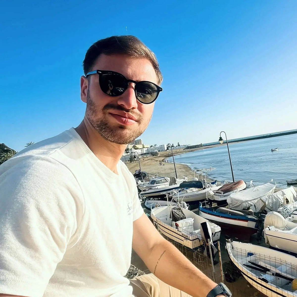 Hombre con camiseta blanca y gafas de sol redondas de pasta negra con lentes verdes frente a barcos en la playa.