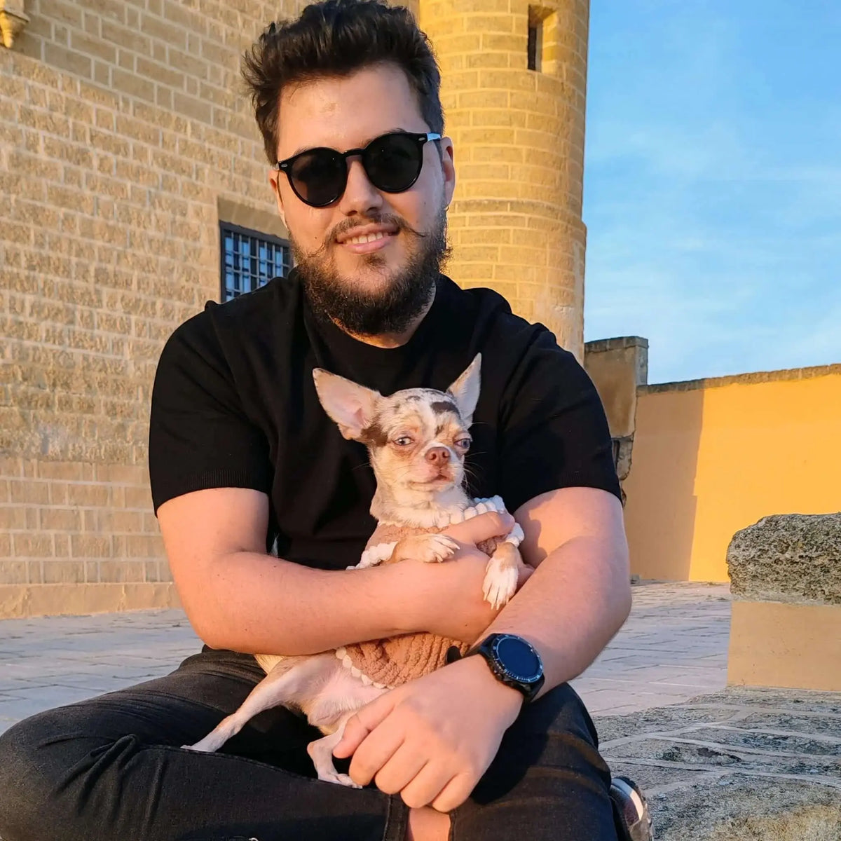 Hombre con gafas de sol negras de montura redonda y lentes verdes oscuras abrazando a un perro frente a un castillo.