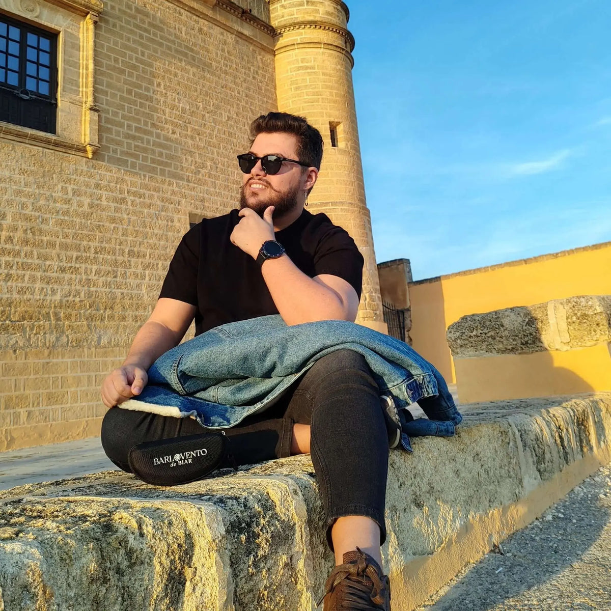 Hombre con gafas de sol negras y lentes verdes polarizadas, estilo casual, sentado ante un castillo de piedra.