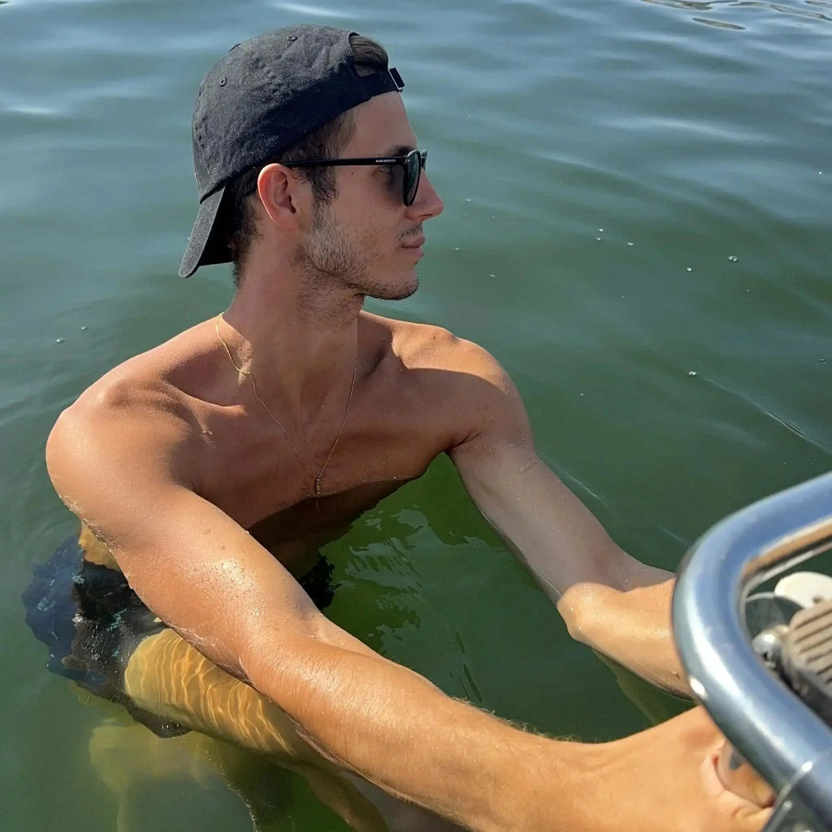 Hombre con gorra negra y gafas de sol de montura negra y lentes polarizadas verdes nadando en el mar.