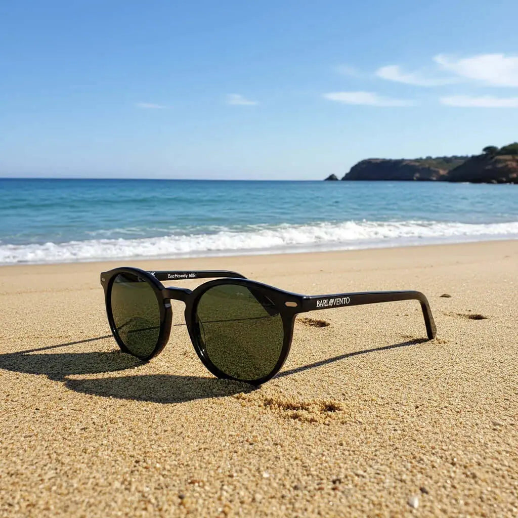 Gafas de sol negras de estilo clásico con lentes polarizadas verdes en la arena de una playa con el mar de fondo.