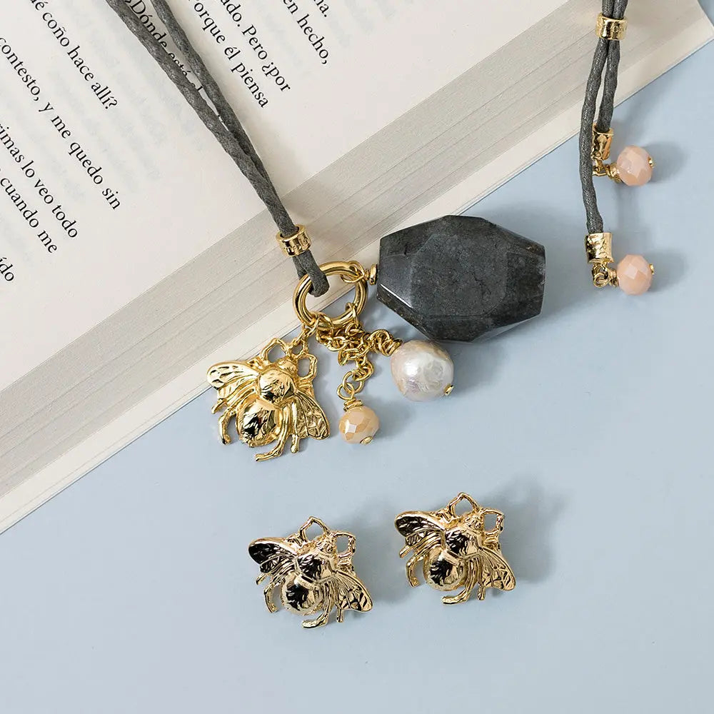 Collar gris con colgante negro, cuentas de perla y aretes dorados con diseño de abeja.