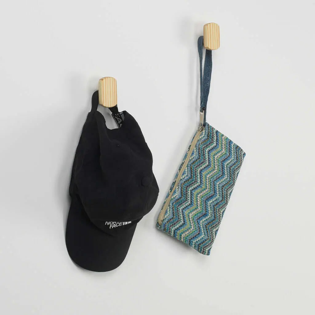 Dos colgadores de pared en madera clara con una gorra negra y un estuche estampado colgados sobre fondo blanco.