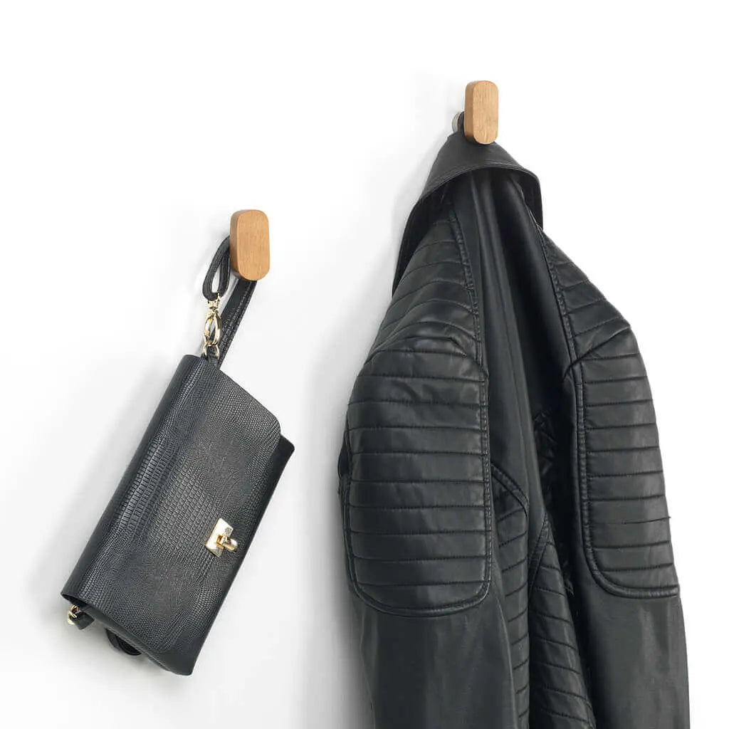 Dos perchas de pared en madera clara con forma ovalada, sosteniendo un bolso y una chaqueta de cuero negra.