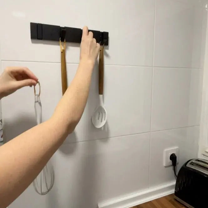 Perchero de pared metálico negro, moderno y minimalista, instalado en azulejos blancos con utensilios colgados.