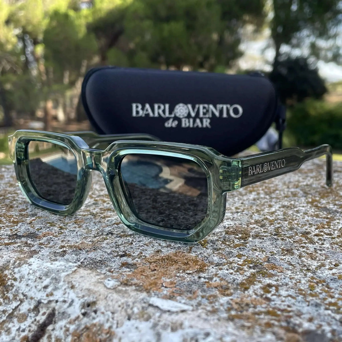 Gafas de sol de montura cuadrada en acetato translúcido color verde y lentes oscuras, sobre una superficie de piedra.