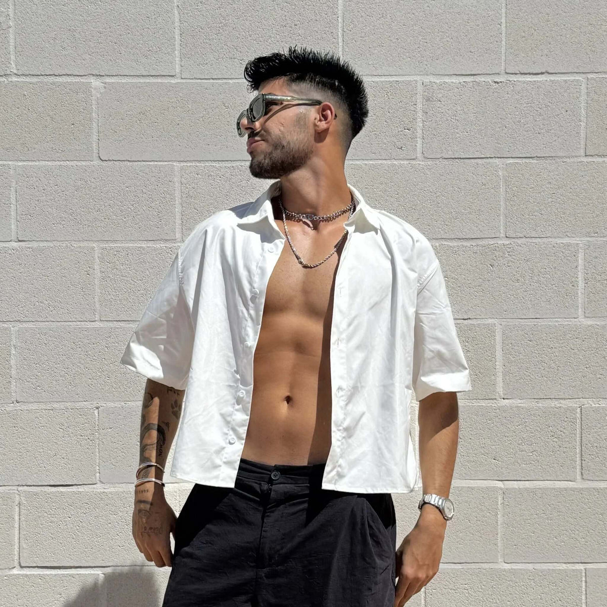 Gafas de sol con montura verde y lentes negras, estilo casual con camisa blanca abierta sobre pared de ladrillo.