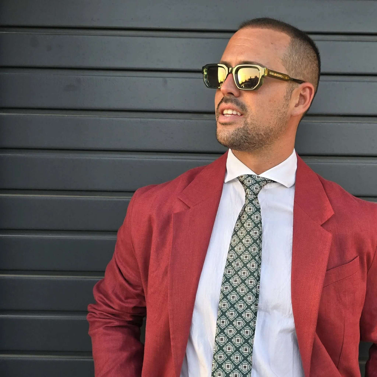 Hombre con gafas de sol de montura verde, lentes oscuras reflectantes, camisa blanca, corbata estampada y blazer rojo.