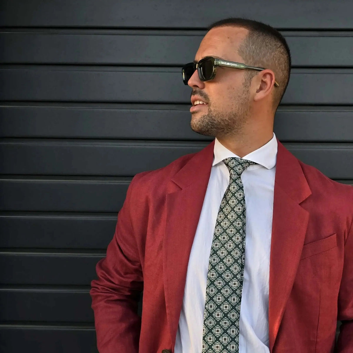 Gafas de sol con montura verde estilo carey y lentes negras oscuras, puestas en un hombre con americana roja.