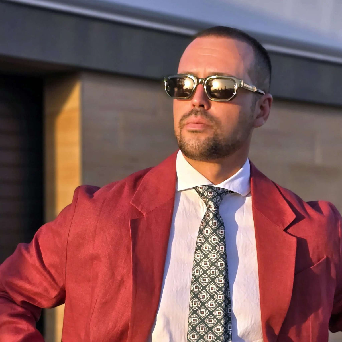 Hombre con gafas de sol de montura verde translúcida y lentes oscuras, traje rojo y corbata estampada.