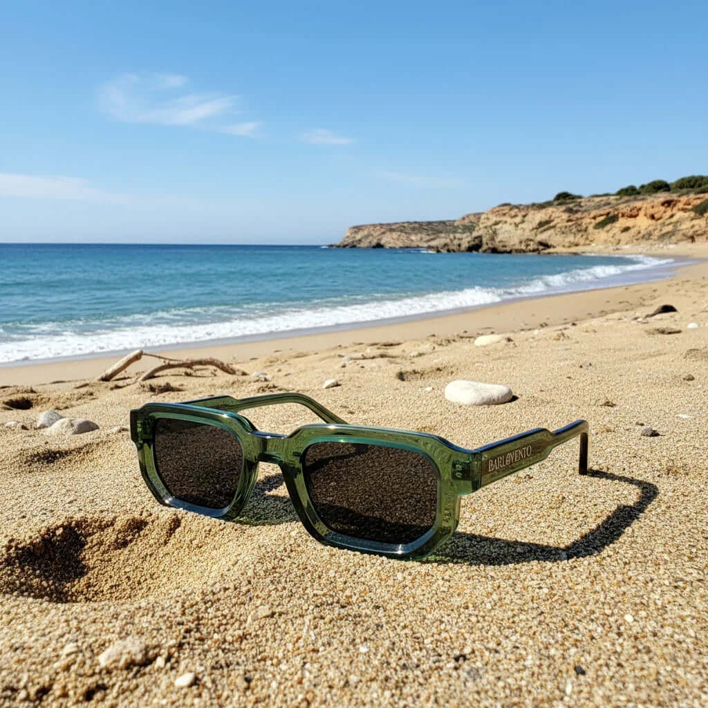 Gafas de sol rectangulares con montura de acetato verde translúcido y lentes negras sobre la arena de la playa.
