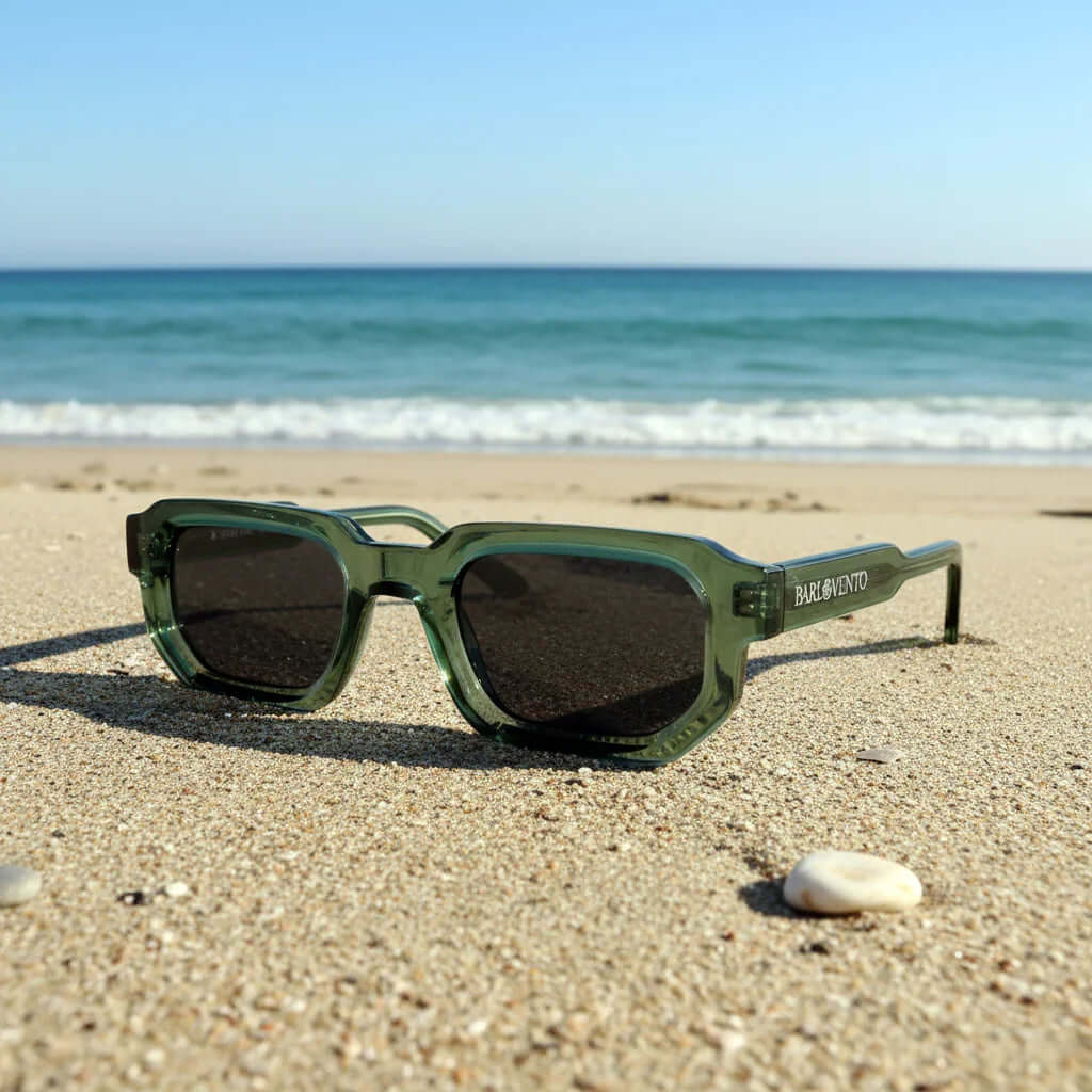 Gafas de sol de montura verde translúcida y diseño geométrico con lentes oscuras sobre la arena de la playa.