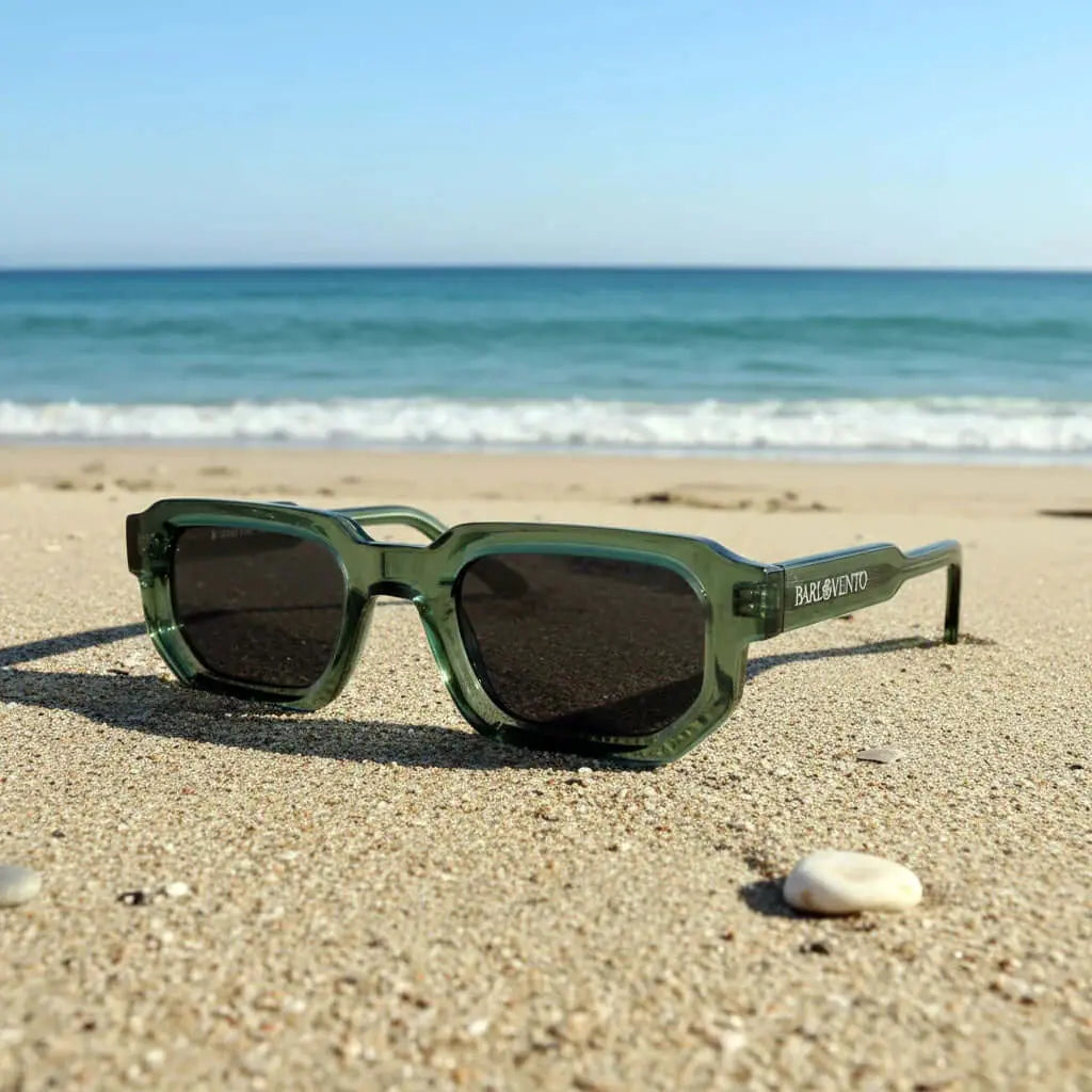 Gafas de sol de montura verde translúcida y diseño geométrico con lentes oscuras sobre la arena de la playa.