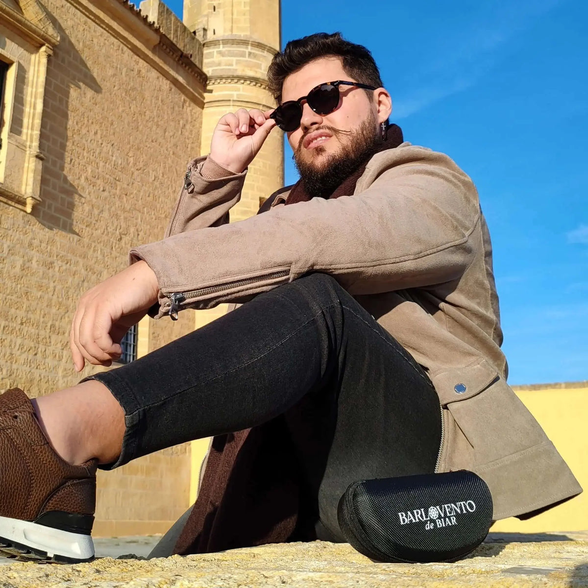 Hombre con gafas de sol redondas de carey y lentes marrones polarizadas, vistiendo chaqueta beige frente a un castillo.