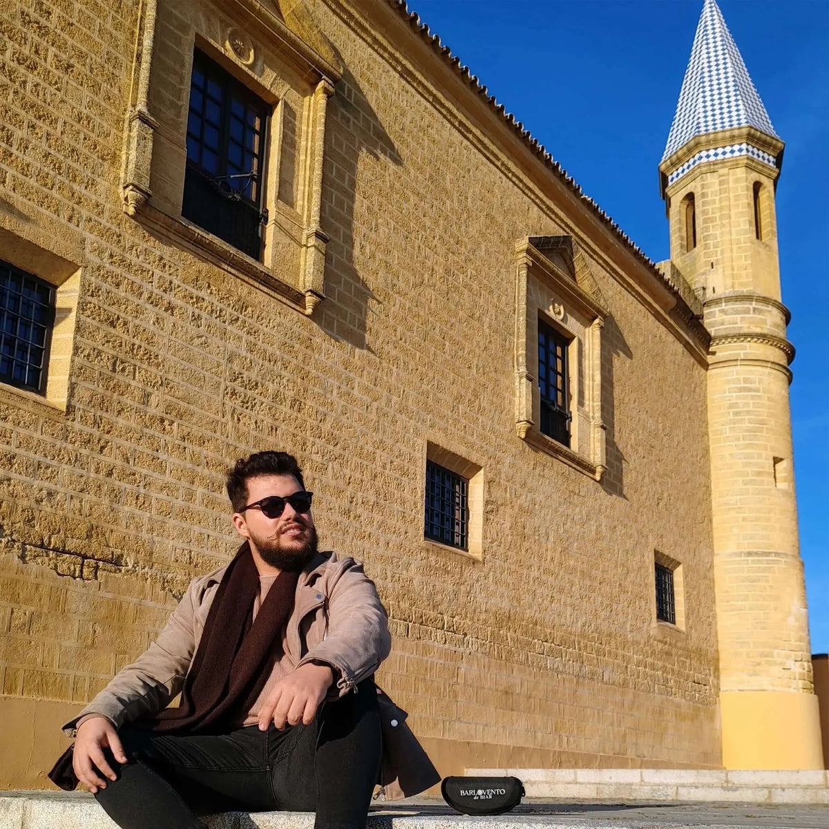 Hombre con gafas de sol de carey y lentes polarizadas marrones, posando frente a un edificio histórico de piedra.