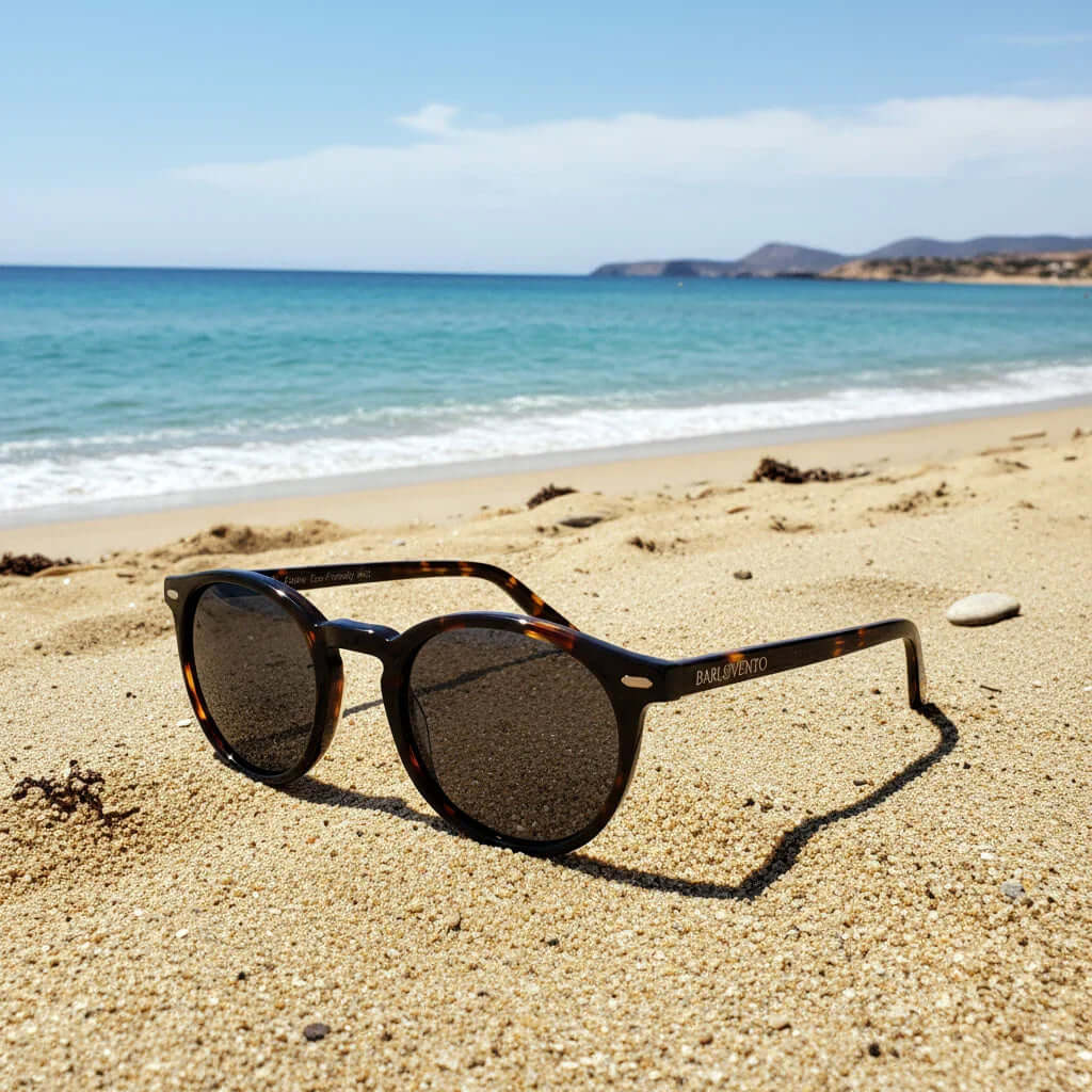Gafas de sol redondas de carey con lentes polarizadas marrones sobre la arena de la playa con el mar de fondo.