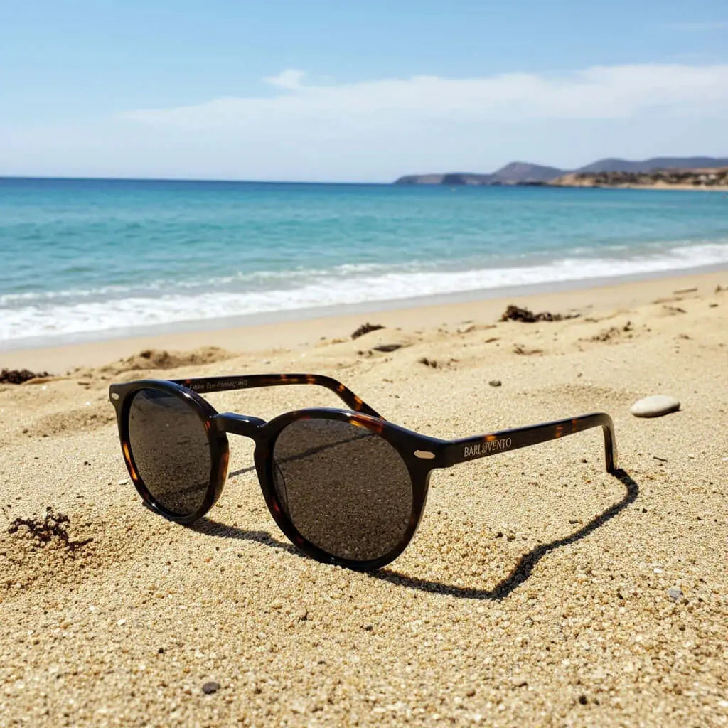 Gafas de sol redondas de carey con lentes polarizadas marrones sobre la arena de la playa con el mar de fondo.