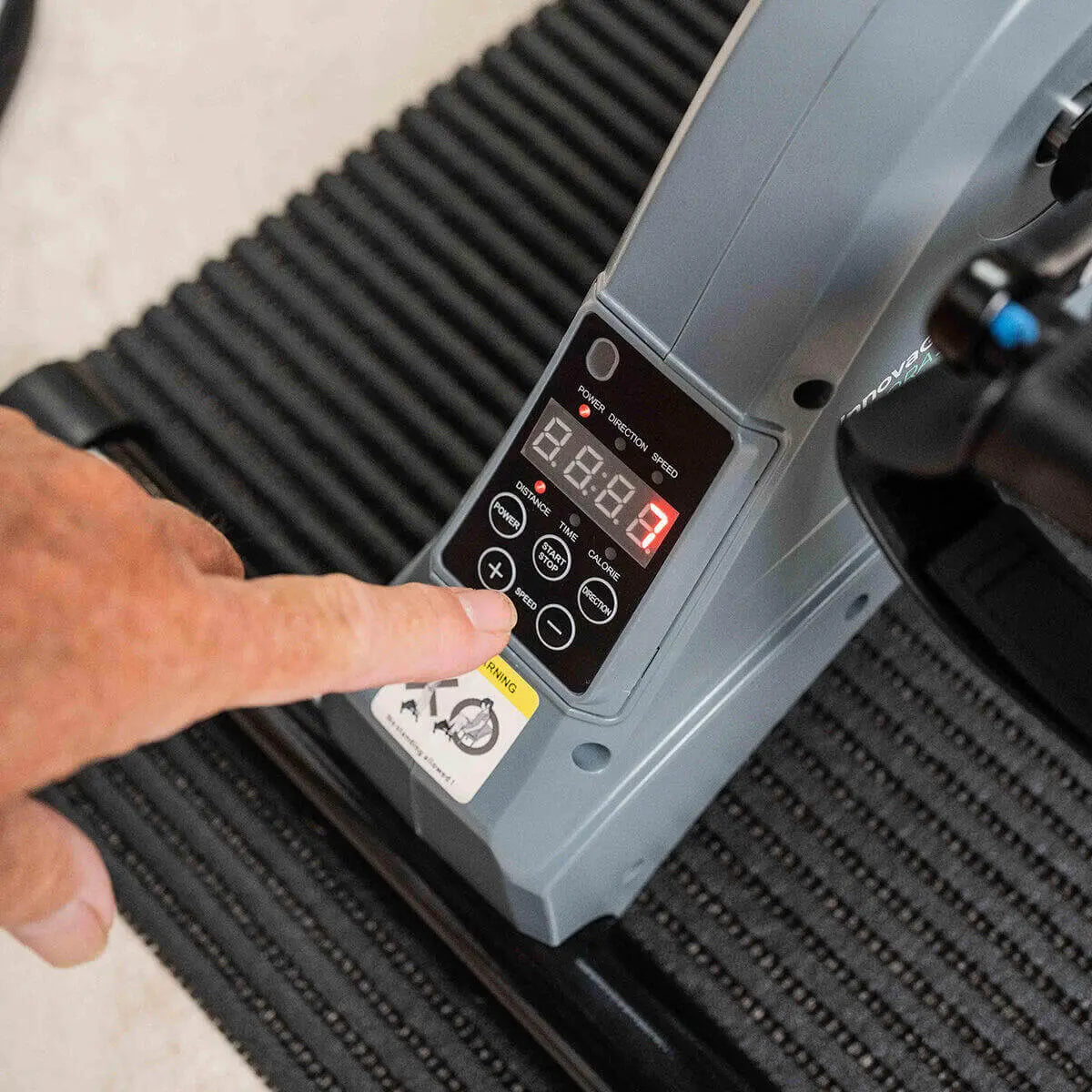Pedaleador eléctrico gris con pantalla digital y botones de control sobre una alfombrilla de goma negra estriada.