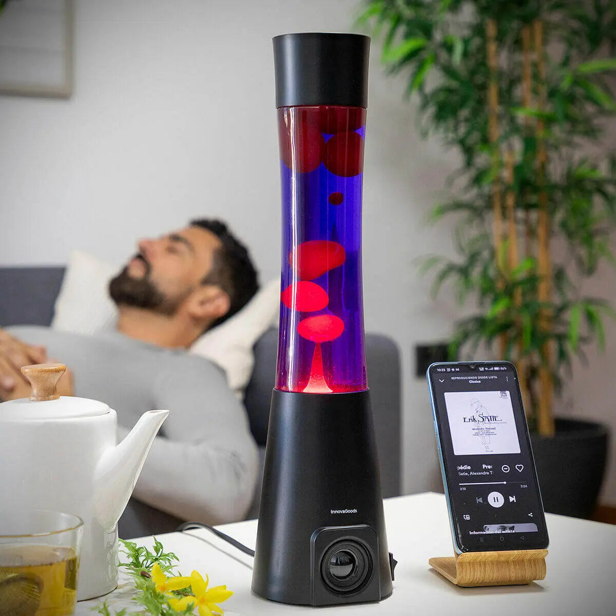 Lámpara de lava cilíndrica con cera roja y líquido violeta sobre base negra con altavoz integrado, junto a un smartphone.