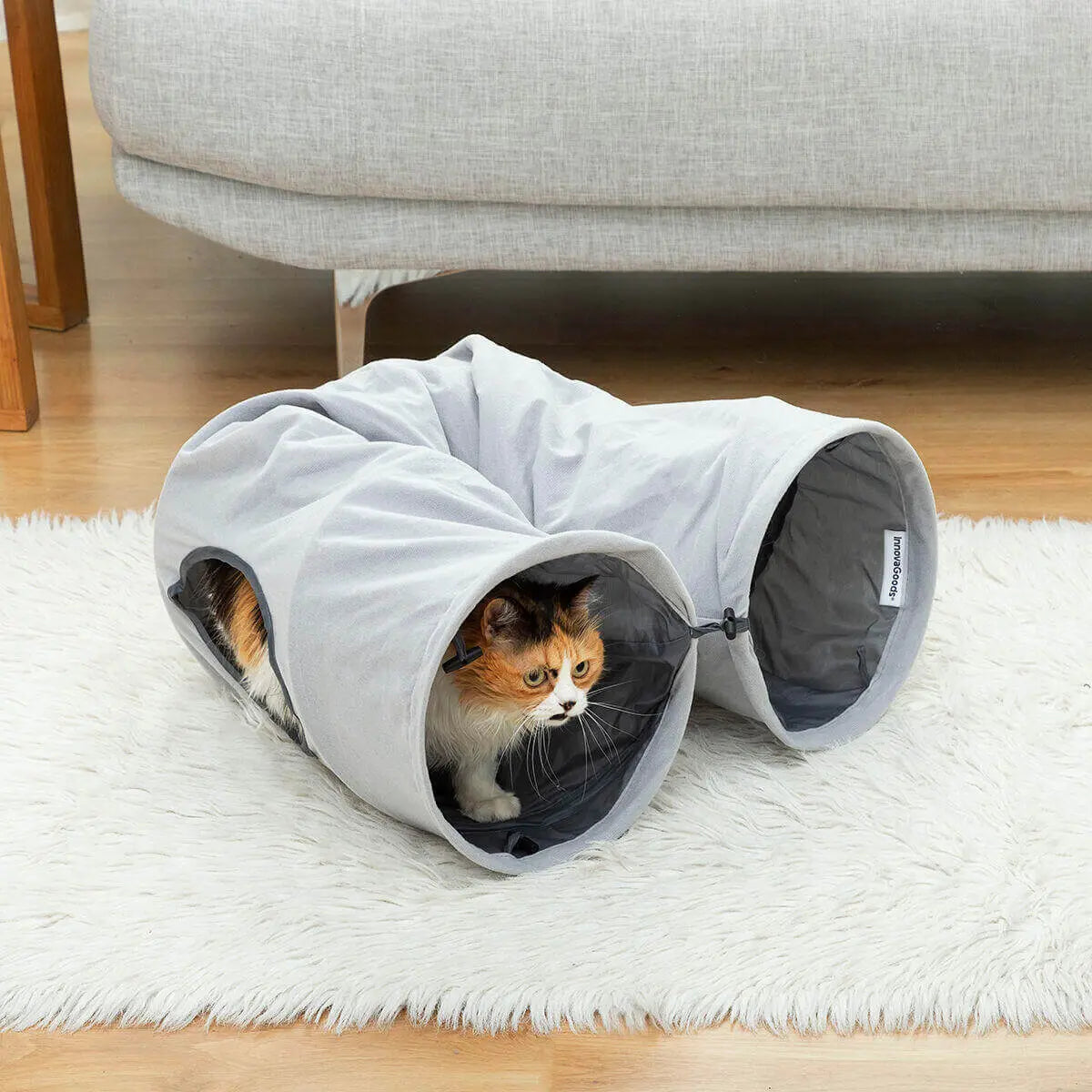 Gato calicó saliendo de un túnel plegable gris de tela suave sobre una alfombra blanca en una sala.