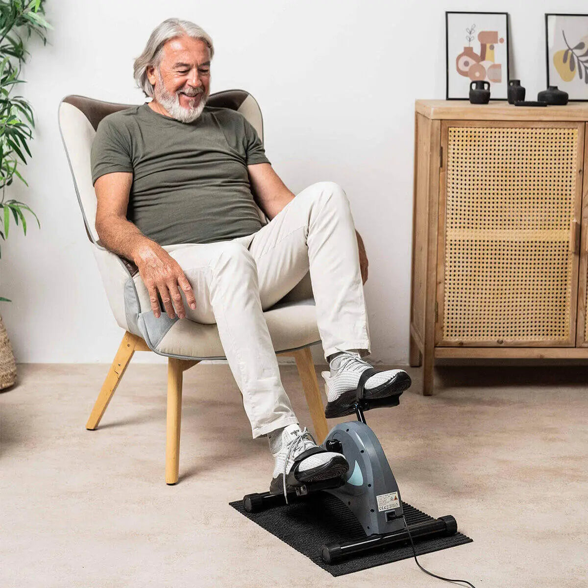 Hombre mayor haciendo ejercicio con un pedaleador eléctrico gris sobre una alfombrilla negra en casa.