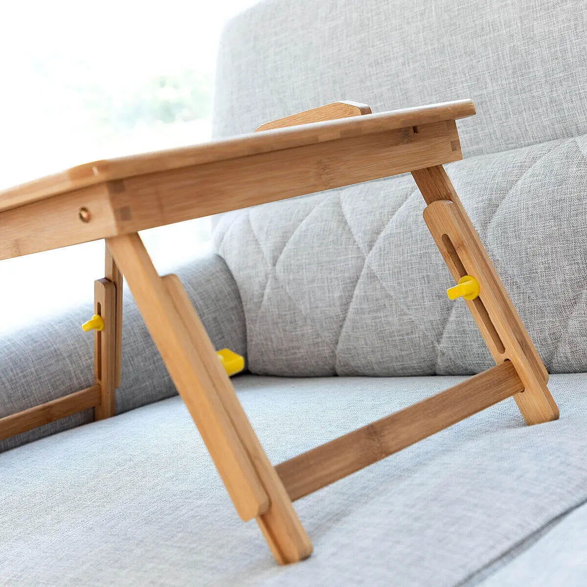 Mesa auxiliar plegable de bambú con altura ajustable y seguros amarillos sobre un sofá gris.