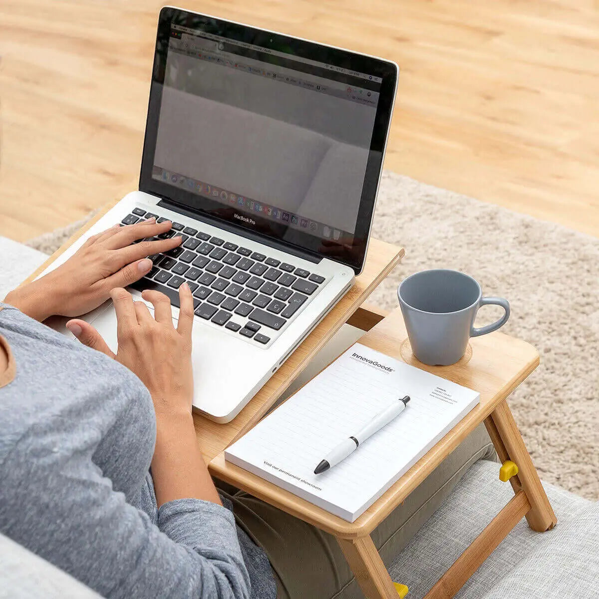 Mesa plegable de bambú sobre las piernas, con portátil, libreta y taza, ideal para trabajar cómodamente en el sofá.