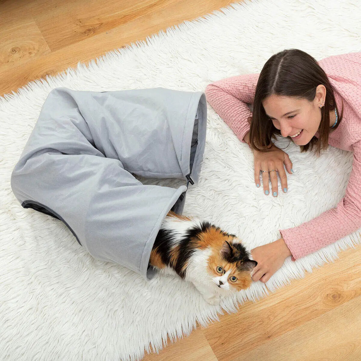 Mujer jugando con un gato tricolor que sale de un túnel gris plegable de tela sobre una alfombra blanca.