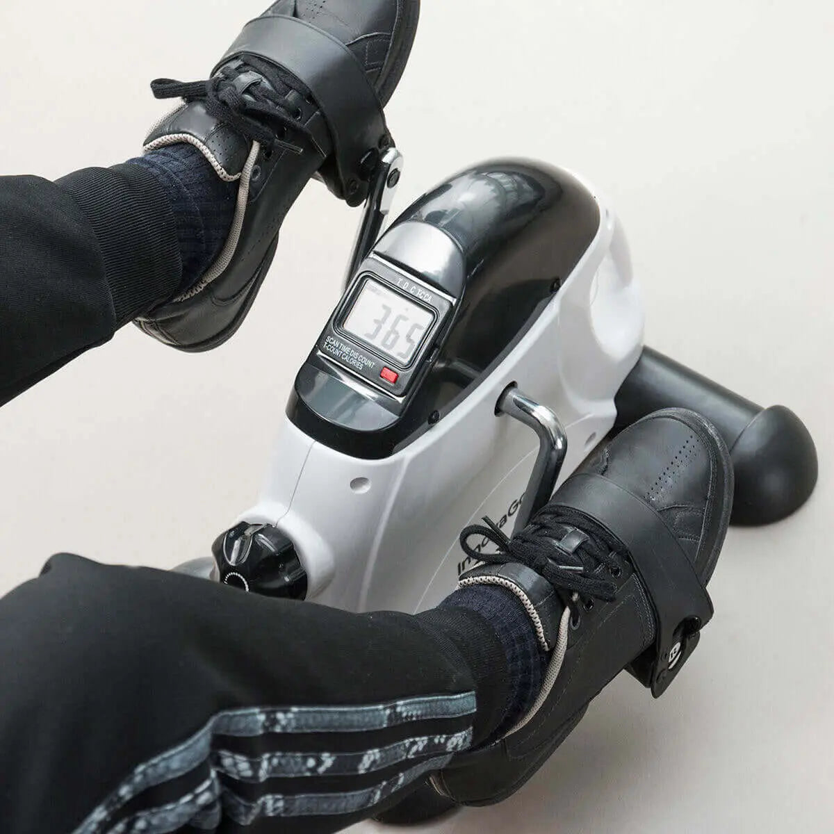 Pedaleador portátil blanco y negro con pantalla LCD, usado con zapatos en los pedales con correas ajustables.